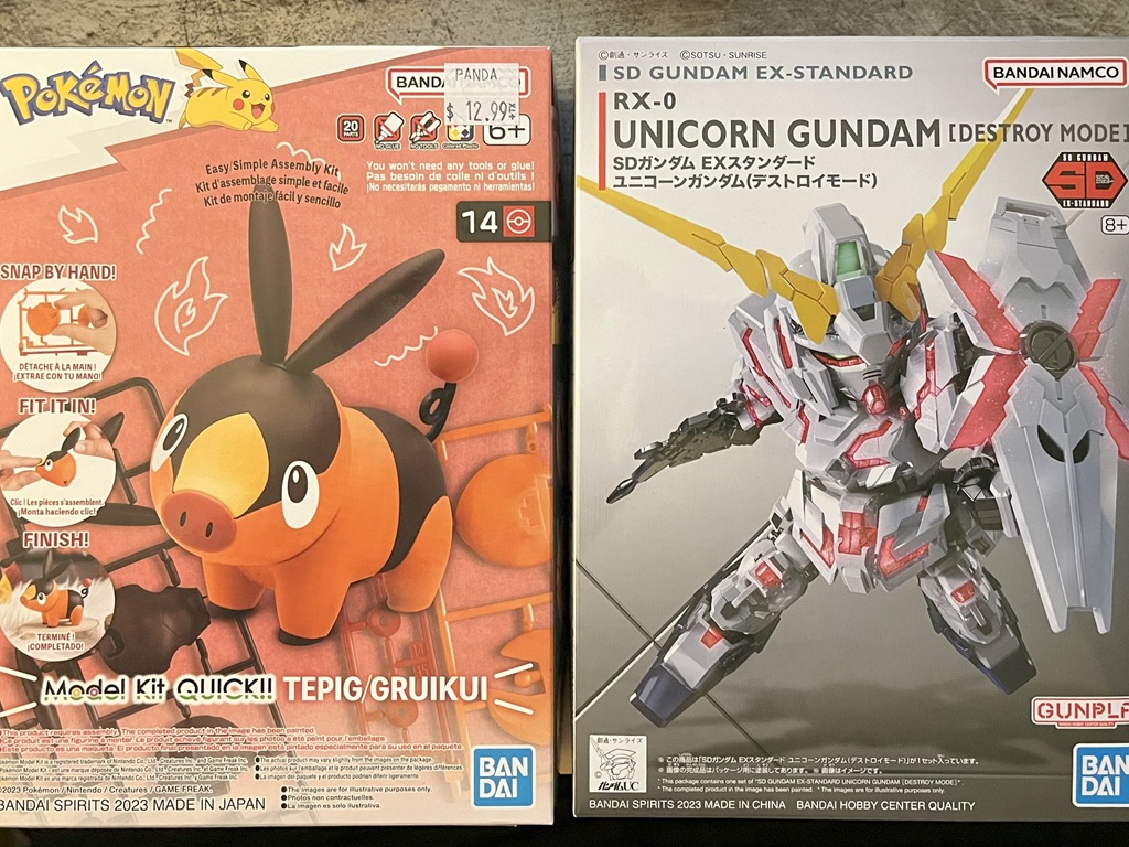 Two plastic model kits. On the left is a model kit for the Pokemon named Tepig, an orange and brown-coloured pig. On the right is a model kit for a Unicorn Gundam mech, which has pink accents and is holding a shield.